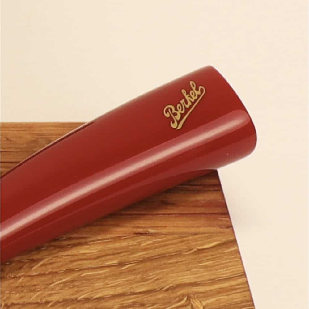 Nahaufnahme eines roten Berkel-Messergriffs, der auf einer hellen Holzoberfläche ruht. Das Wort „Berkel“ ist in goldfarbener Schrift auf dem Griff geschrieben und erinnert an die Präzision eines Elegance Santoku-Messers.