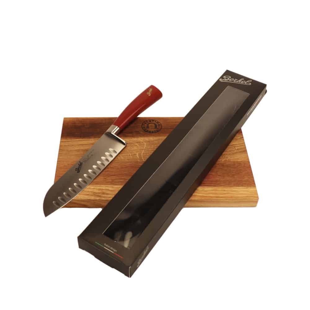 Ein Santoku-Messer mit rotem Holzgriff ruht auf einem 100 % handgefertigten Schneidebrett Unikat mit den Maßen 30 x 18 cm. Das Berkel Messer - Elegance Santoku-Messer verfügt über eine 18 cm lange gezahnte Stahlklinge. Neben dem Messer befindet sich eine schwarze rechteckige Schachtelverpackung mit transparentem Kunststoff, die einen Teil der Klinge freigibt.