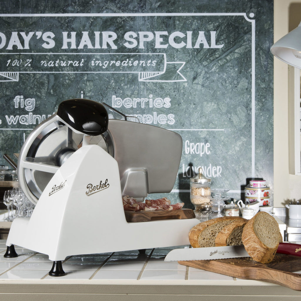 Eine Berkel Red Line 250 Weiss Profi-Aufschnittmaschine (25 cm Messer, Fassholz-Einlegebrett) steht auf der Theke und schneidet Aufschnitt. Daneben liegen ein Brotlaib, ein Brotmesser und Gläser. Im Hintergrund ist eine Kreidetafel zu sehen.