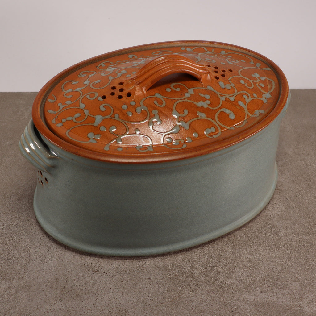 Ein glasierter Brottopf und Vorratstopf, handgefertigt aus Steinzeug, oval, mit einem hellblau-grauen Boden und einem Deckel mit aufwendigen orangefarbenen und blauen Ornamenten. Der Deckel hat einen gebogenen Griff und Entlüftungslöcher, während der Boden mit integrierten Seitengriffen ausgestattet ist. Der Steinzeug-Bräter steht auf einer grauen Oberfläche.