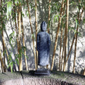 Eine Steinstatue einer stehenden Buddha-Figur aus Lavastein, 65 cm – Ein Kunstwerk voller Weisheit – Harmonie für Körper Seele Haus & Garten steht auf einem Steinsims. Hinter der Statue werfen hohe Bambuspflanzen Schatten auf einen hellen Hintergrund. Die Szene mit ihrer Verwendung von Lavastein und natürlichen Elementen strahlt eine heitere und friedliche Atmosphäre voller Harmonie aus.