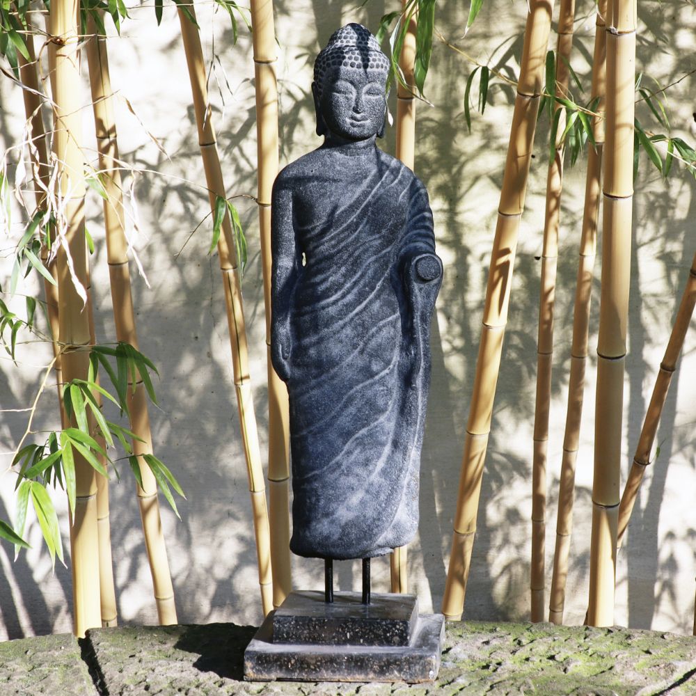 Die stehende Buddha-Figur aus Lavastein (65 cm), ein Meisterwerk voller Weisheit, steht anmutig auf einem Sockel, umgeben von Bambusstäben und -blättern. In ihrer ruhigen, aufrechten Haltung mit traditioneller drapierter Kleidung verkörpert die Statue Harmonie in dieser friedlichen und natürlichen Umgebung und ist damit eine ideale Ergänzung für Körper, Seele, Haus und Garten.