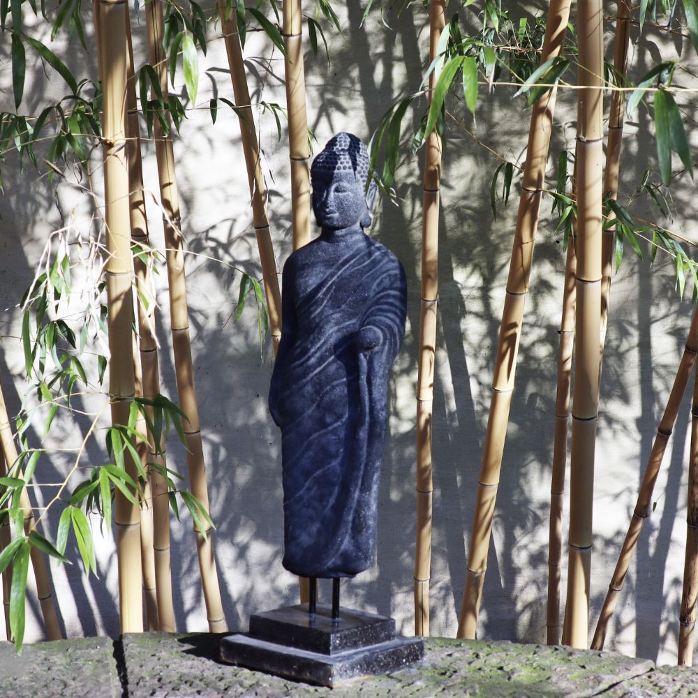 Eine dunkle stehende Buddha-Figur aus Lavastein (65 cm) - Ein Kunstwerk voller Weisheit, die auf einem rechteckigen Sockel steht, steht vor einem Hintergrund aus hohen, dünnen Bambuspflanzen. Das Lavastein-Kunstwerk, teilweise im Schatten, scheint in einer ruhigen Außenumgebung mit einer verwitterten Wand dahinter zu stehen.