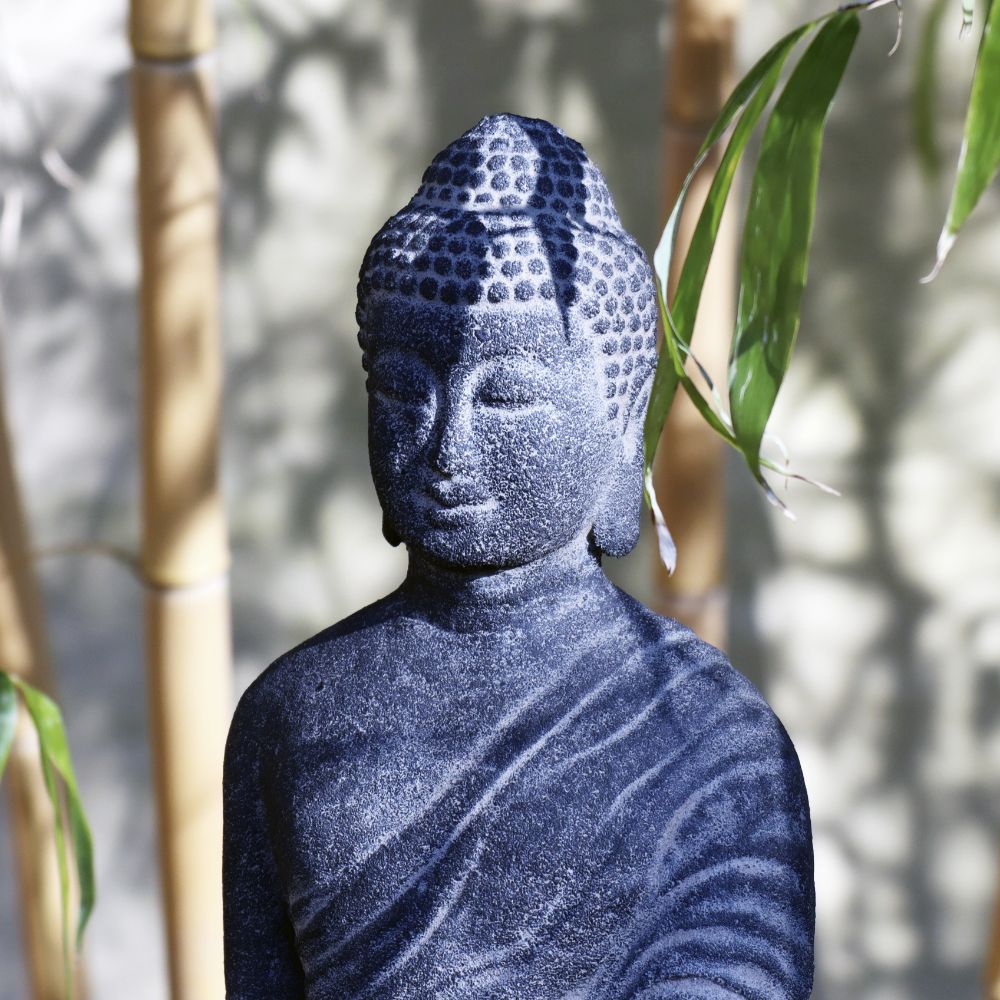 Eine stehende Buddhafigur aus Lavastein, 65 cm groß und die reine Weisheit verkörpernd, steht gelassen zwischen Bambusstäben und -blättern. Diese Steinstatue stellt Buddha mit friedlichem Ausdruck dar und ist mit traditionellen Gewändern geschmückt. Sanftes Licht wirft sanfte Schatten, die die Texturen und Merkmale hervorheben und Harmonie für Körper Seele Haus & Garten schaffen.
