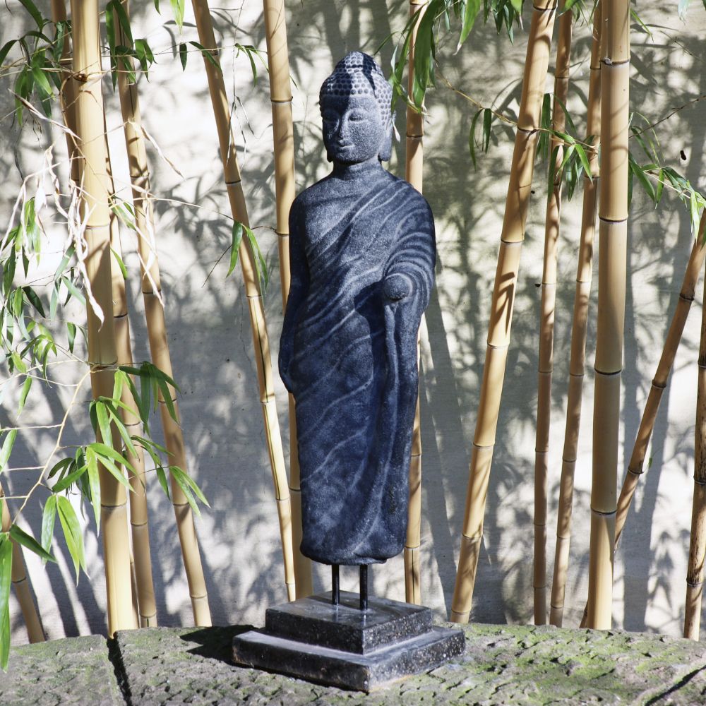 Eine stehende Buddhafigur aus Lavastein, 65 cm, ein Kunstwerk voller Weisheit, steht gelassen da und hat eine Hand in einer Segensgeste erhoben. Die Statue steht auf einem kleinen Sockel und ist von hohen Bambusstäben umgeben, die eine friedliche, Zen-artige Atmosphäre schaffen. Sonnenlicht fällt durch die Bambusblätter und verstärkt das Gefühl der Harmonie für Körper, Seele, Haus und Garten.