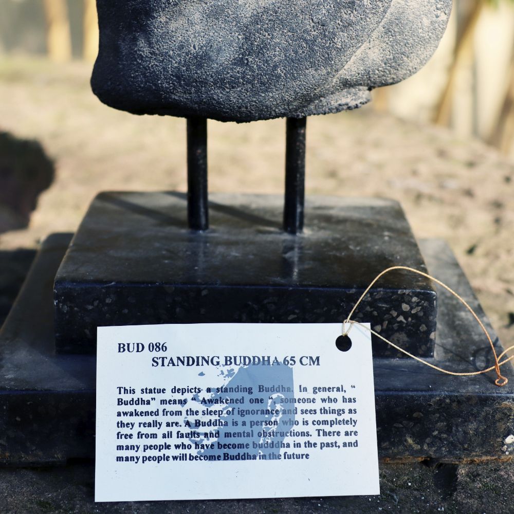 Nahaufnahme des Sockels einer „Stehenden Buddha Figur aus Lavastein 65 cm – Ein Kunstwerk voller Weisheit – Harmonie für Körper Seele Haus & Garten“, mit einem angebrachten Informationsschild. Auf dem Schild steht: „BUD 086 STEHENDER BUDDHA 65 CM“ und beschreibt die Bedeutung der Erleuchtung. Der Hintergrund ist eine verschwommene natürliche Außenumgebung, die Harmonie für Körper, Seele, Haus und Garten verkörpert.