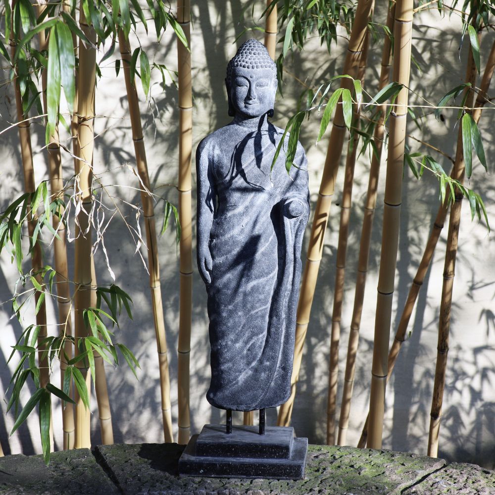 Eine 75 cm große, stehende Buddhafigur aus Lavastein steht inmitten üppiger Bambuspflanzen. Die Statue, ein Kunstwerk voller Weisheit, zeigt Buddha in einer ruhigen Pose mit einer erhobenen Hand und der anderen an seiner Seite. Sonnenlicht fällt durch den Bambus und wirft sanfte Schatten auf diese Lavasteinfigur, die Harmonie für Körper Seele Haus & Garten bringt.