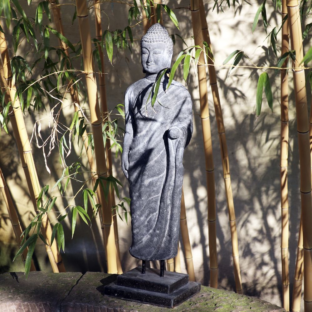 Eine stehende Buddha-Figur aus Lavastein (75 cm) steht anmutig vor einem Hintergrund aus hohen Bambuspflanzen. Das Kunstwerk voller Weisheit zeigt Buddha in einer ruhigen Haltung mit einer erhobenen Hand und der anderen an der Taille. Sonnenlicht fällt durch die Bambusblätter und wirft komplizierte Schatten auf die Lavastein-Skulptur, die Körper, Seele, Haus und Garten in Harmonie bringt.