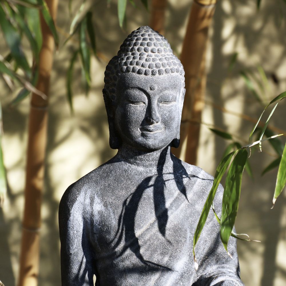 Die „Stehende Buddha Figur aus Lavastein 75cm“, ein Kunstwerk voller Weisheit, steht anmutig zwischen Bambusstäben und Blättern und strahlt einen ruhigen Ausdruck aus. Sonnenlicht fällt durch, wirft Schatten auf die Lavasteinskulptur und schafft eine heitere, harmonische Atmosphäre für Körper und Seele in Ihrem Haus oder Garten.