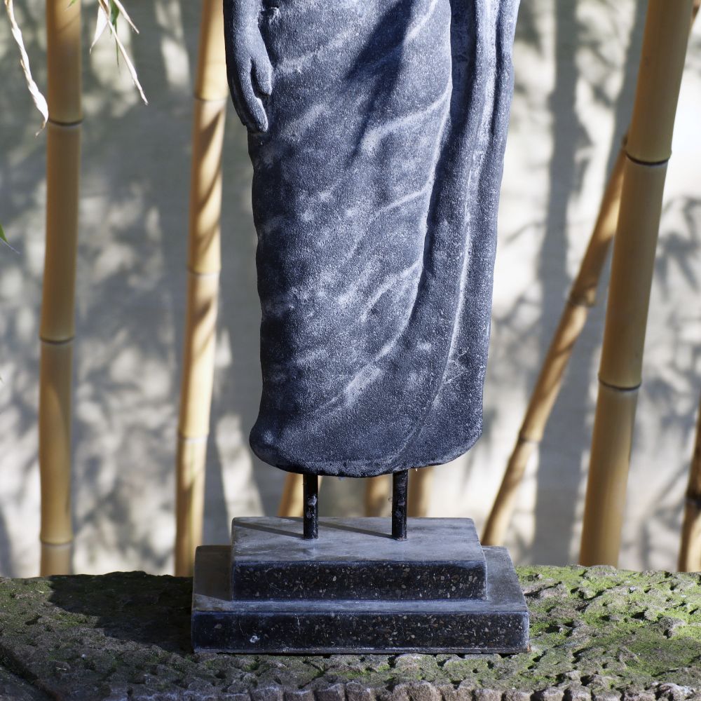 Eine Nahaufnahme einer Steinstatue „Stehende Buddha-Figur aus Lavastein 75 cm – Ein Kunstwerk voller Weisheit – Harmonie für Körper Seele Haus & Garten“ von der Hüfte abwärts zeigt drapierte Kleidung. Die 75 cm große Statue steht auf einem kleinen Sockel, der von einer strukturierten Steinoberfläche umgeben ist. Im unscharfen Hintergrund sind Bambusstäbe zu sehen, die auf eine Außenumgebung mit Lavastein-Akzenten hindeuten.