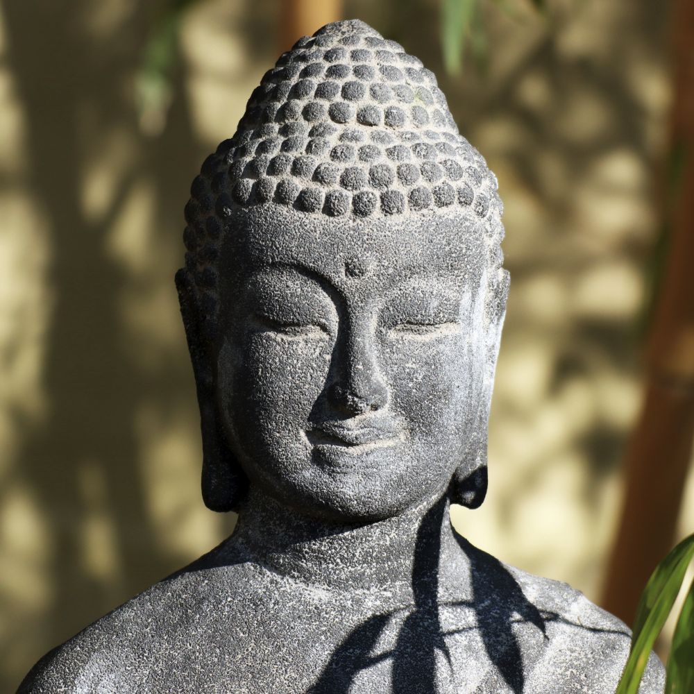 Eine Steinstatue von Buddha mit ruhigem Gesichtsausdruck, geschlossenen Augen und einem heiteren Lächeln. Die stehende Buddha-Figur aus Lavastein (75 cm) ist detailliert mit strukturiertem Haar und glatten Gesichtszügen. Das Sonnenlicht wirft Schatten, die das Gefühl von Harmonie für Körper Seele Haus & Garten inmitten der verschwommenen grünen Pflanzen im Hintergrund verstärken.