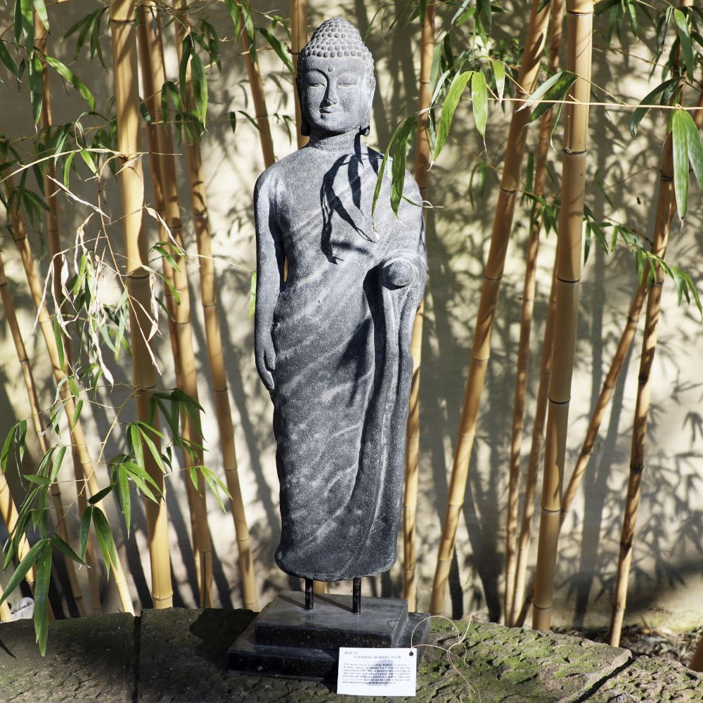 Eine „Stehende Buddha Figur aus Lavastein 75cm – Ein Kunstwerk voller Weisheit“ ist anmutig von Bambuspflanzen umgeben. Die detaillierte, in Gewänder gehüllte Statue steht draußen auf einem schwarzen Sockel. Sonnenlicht erzeugt sowohl auf der Statue als auch auf dem Bambus komplizierte Schatten und verkörpert Harmonie für Körper Seele Haus & Garten.