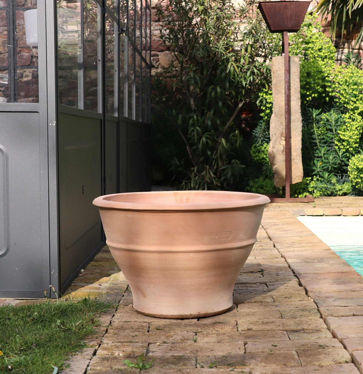 Ein Terrakotta-Pflanzgefäß „Buxus“, 50 cm, steht auf einem mit Steinplatten ausgelegten Weg neben einem Gewächshaus und einem Swimmingpool, umgeben von grünen Blattpflanzen und Buchsbäumen. Das Pflanzgefäß ist leer und prominent im Vordergrund des Gartenbereichs platziert.