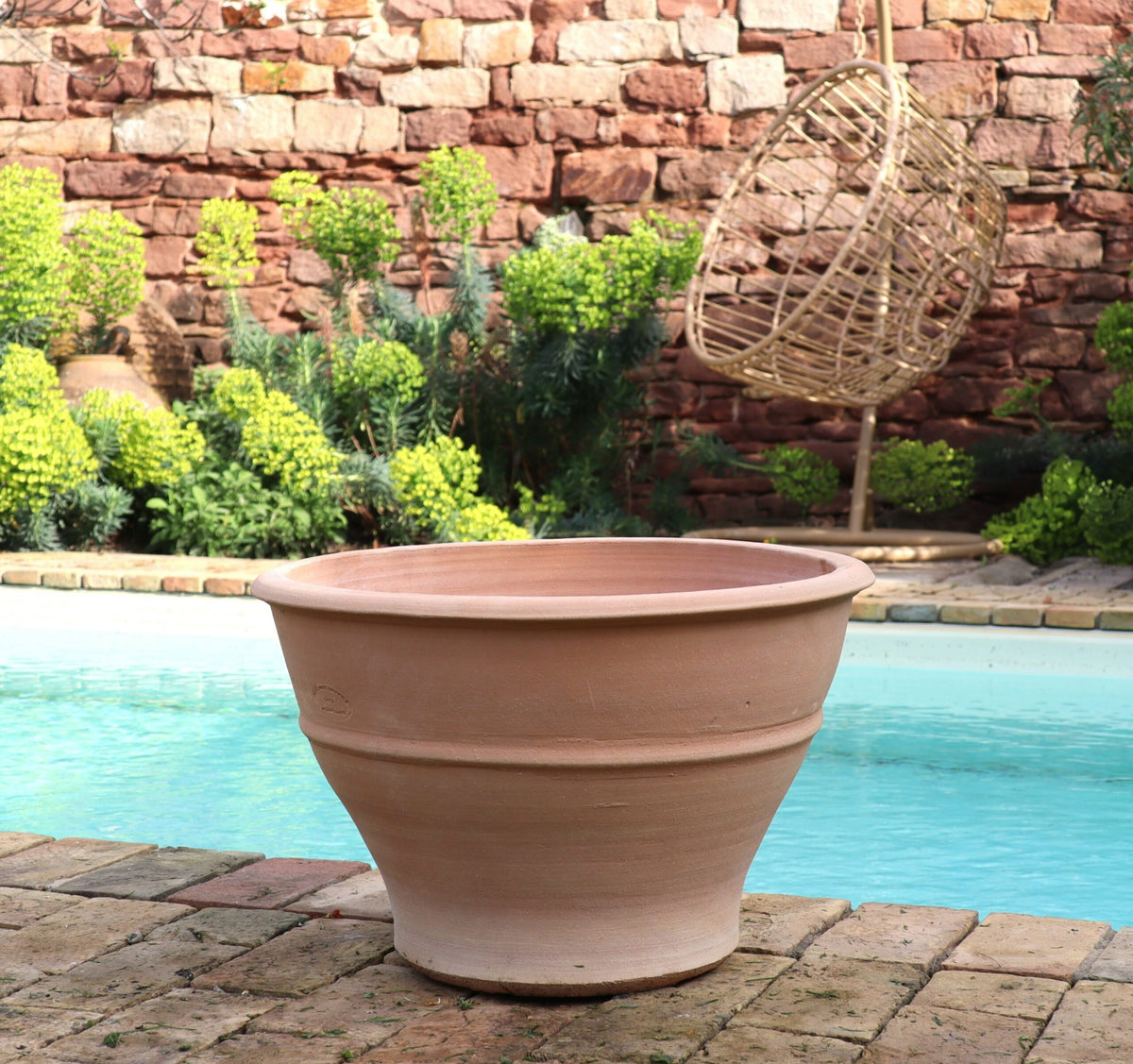 Ein großes, leeres Terrakotta-Pflanzgefäß „Buxus“, 50 cm, steht auf einer gemauerten Terrasse neben einem Swimmingpool. Im Hintergrund sind eine Steinmauer und üppiges Grün zu sehen, auf der rechten Seite hängt ein hängender Korbstuhl.