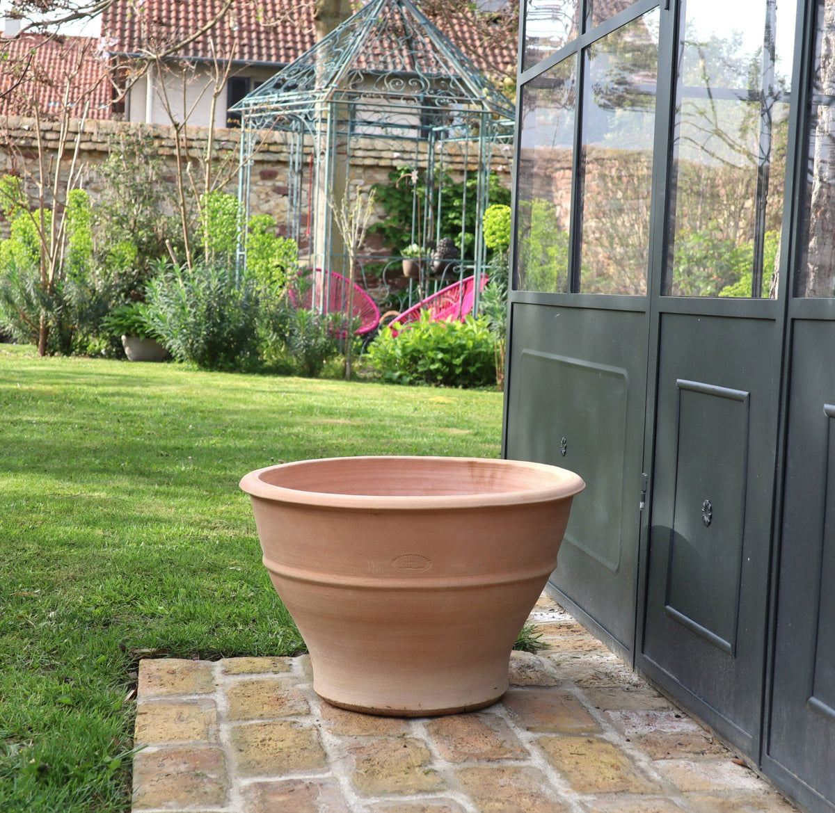 Ein 50 cm großes Pflanzgefäß aus Terrakotta „Buxus“ steht auf einer mit Ziegeln gepflasterten Fläche neben einer dunkel gefärbten Metall- und Glaskonstruktion, möglicherweise einem Gewächshaus. Im Hintergrund ist ein Garten mit grünem Gras, Buxus-Sträuchern, einem Metallpavillon und einem Haus mit Ziegeldach zu sehen.