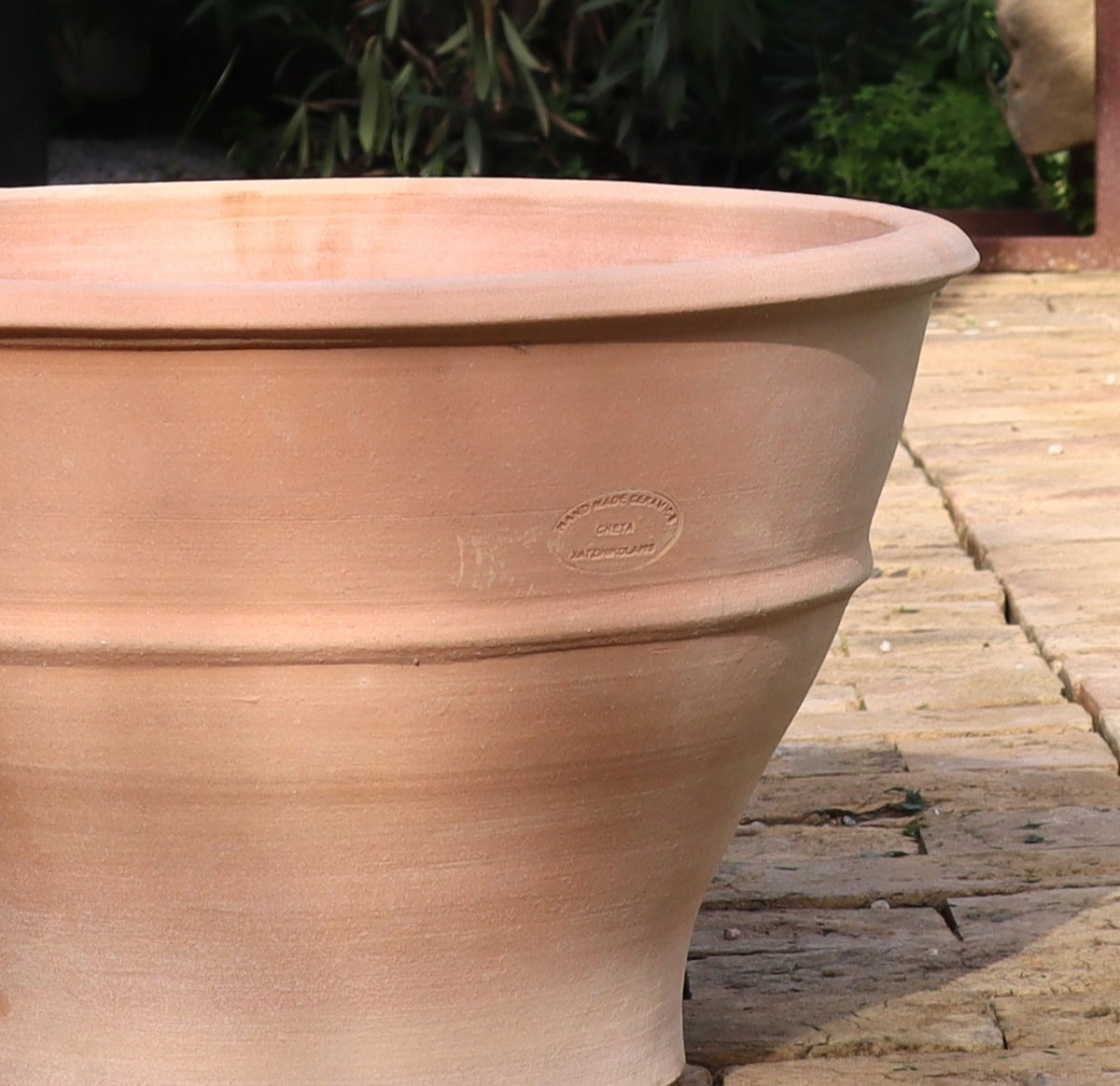 Ein Terrakotta-Pflanzgefäß „Buxus“, 50 cm mit Herstellermarke steht auf einer mit Steinfliesen ausgelegten Terrasse. Das Pflanzgefäß weist ein schlichtes, konisches Design auf und ist von grünem Blattwerk und Buxuspflanzen im Hintergrund umgeben.