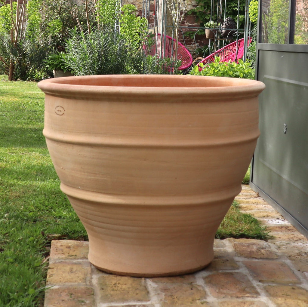 Ein großer, leerer Terrakotta-Pflanztopf namens winterhartes Pflanzgefäß „Buxus“, 60 cm steht auf einem gepflasterten Bereich in einem üppigen Garten. Er verfügt über horizontale Rillen und einen breiten Rand, der Buxus-Sträucher perfekt in Szene setzt. Im Hintergrund sind Grünpflanzen und moderne rosafarbene Gartenstühle zu sehen.