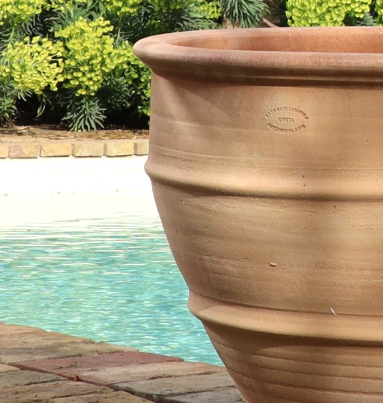 Ein großes winterhartes Pflanzgefäß „Buxus“ mit 60 cm Durchmesser steht leer auf einer Steinterrasse neben einem Swimmingpool. Im Hintergrund sind üppige grüne Blätter zu sehen, darunter ordentlich geschnittene Buxus. Die Szene ist hell vom Sonnenlicht erhellt, was die ruhige Außenumgebung und den robusten Pflanztopf hervorhebt.