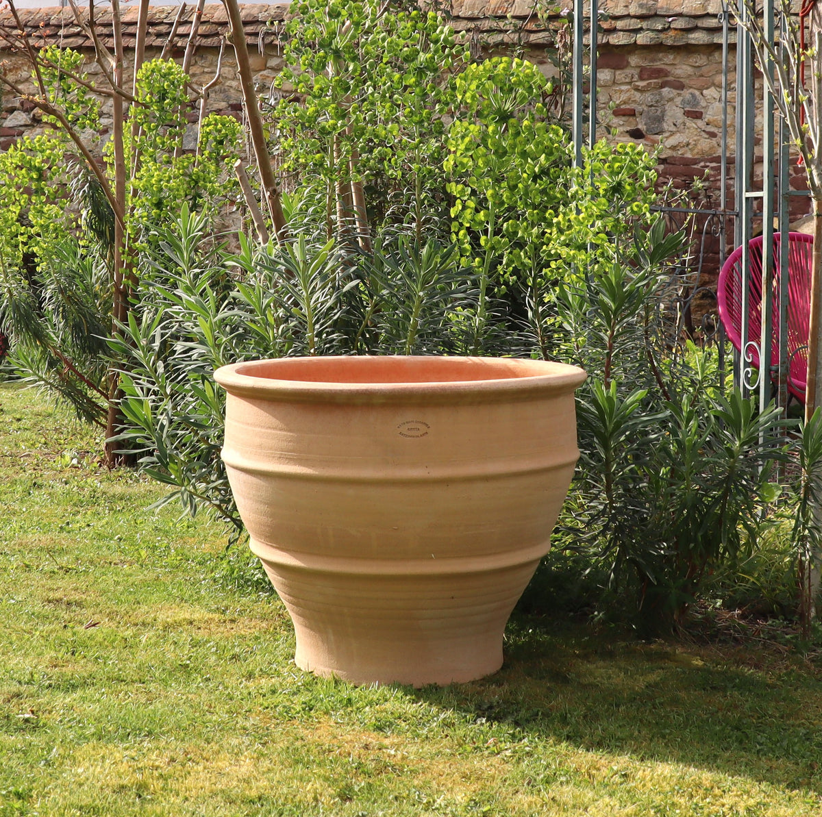 Ein großes, beiges winterhartes Pflanzgefäß „Buxus“, 60 cm im Durchmesser, steht auf einer Rasenfläche. Im Hintergrund umgeben verschiedene Grünpflanzen und Sträucher Teile einer Backsteinmauer und einer metallenen Gartenstruktur. Die Szenerie erinnert an einen gepflegten Garten.