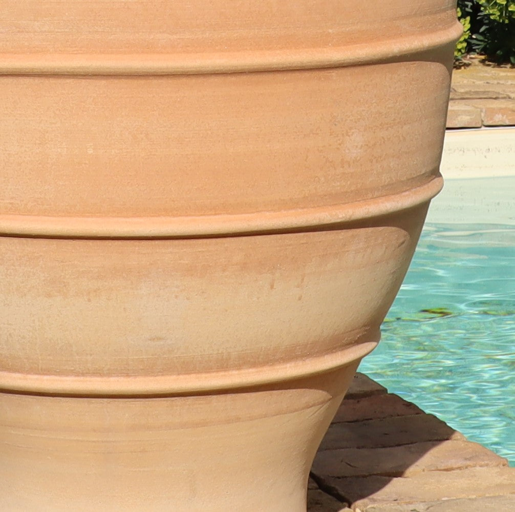 Ein großer, terrakottafarbener frostfester XXL Pflanzkübel „Buxus“, 70 cm mit waagerechten Rillen steht auf einer gefliesten Außenfläche neben einem Swimmingpool. Das Wasser im Pool wirkt ruhig und reflektiert das Sonnenlicht.