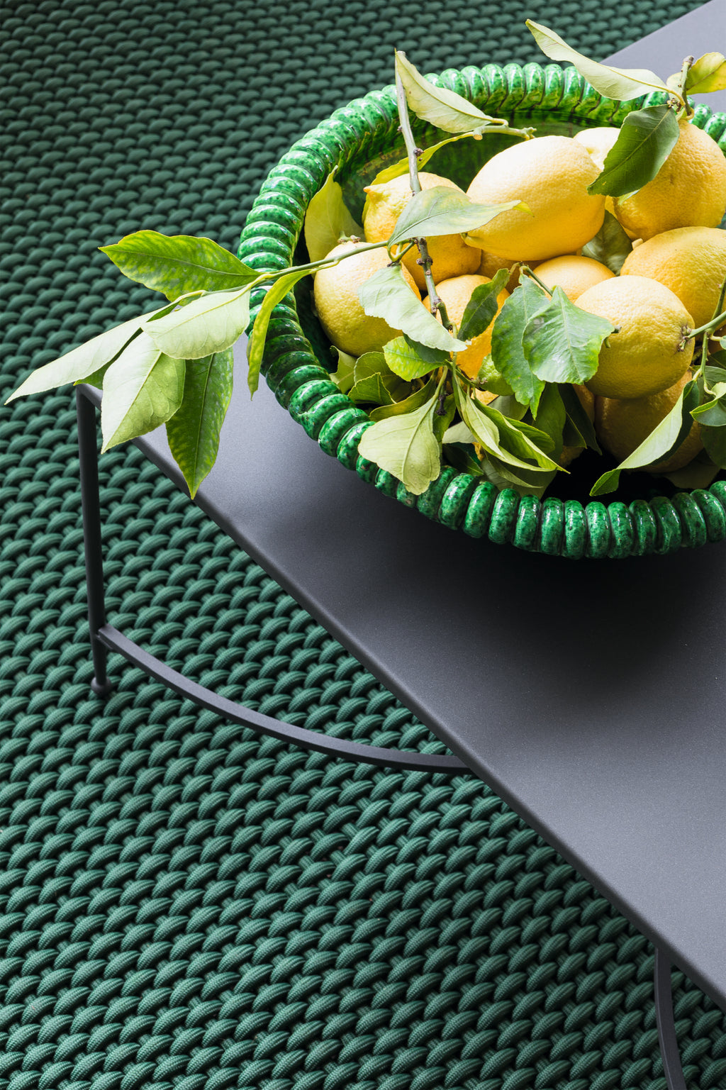 Ein grüner Korb voller frischer Zitronen und Zitronenblätter steht auf einem dunklen, modernen graphitgrauen Metalltisch. Der Tisch steht auf einer strukturierten, grün gewebten Oberfläche, die den Korb und die Zitronen ergänzt. Die Gesamtästhetik ist lebendig und erfrischend und wertet den Raum auf wie ein Sessel Toscana aus Eisen in einem eleganten Ambiente.