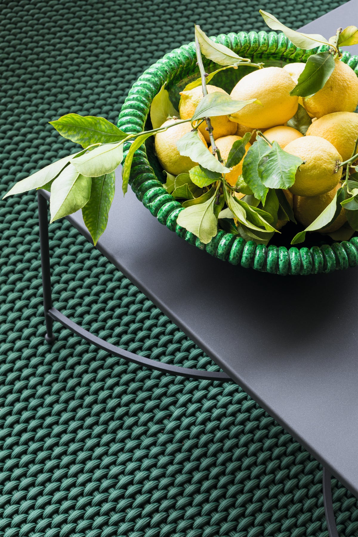 Ein grüner Korb voller frischer Zitronen und Zitronenblätter steht auf einem dunklen, modernen graphitgrauen Metalltisch. Der Tisch steht auf einer strukturierten, grün gewebten Oberfläche, die den Korb und die Zitronen ergänzt. Die Gesamtästhetik ist lebendig und erfrischend und wertet den Raum auf wie ein Sessel Toscana aus Eisen in einem eleganten Ambiente.