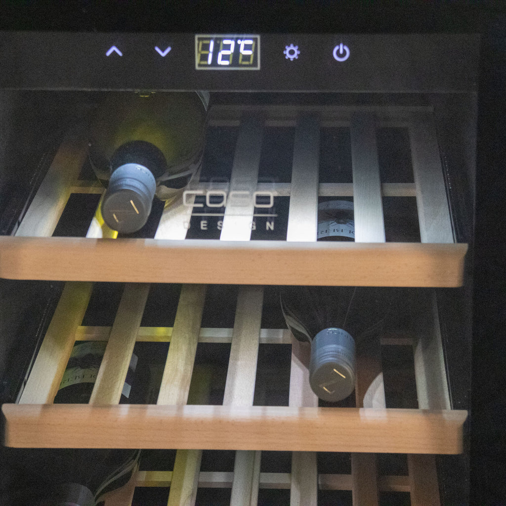 Der Weinkühlschrank 2 Zonen CASO WineComfort 38 in schwarz mit Fassholz-Umhausung ist mit mehreren Weinflaschen gefüllt, die auf Holzregalen ruhen. Die Temperaturanzeige am Kühlschrank zeigt „12°C“. Der Markenname „COCO DESIGN“ ist auf der Glastür zu sehen und entspricht der Qualität, die Sie von diesem CASO WineComfort-Modell erwarten.