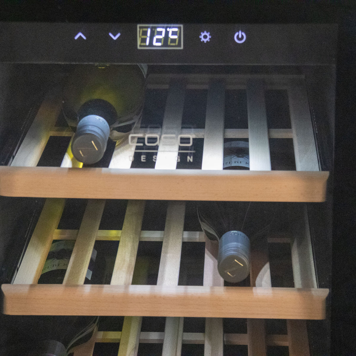 Der Weinkühlschrank 2 Zonen CASO WineComfort 38 in schwarz mit Fassholz-Umhausung ist mit mehreren Weinflaschen gefüllt, die auf Holzregalen ruhen. Die Temperaturanzeige am Kühlschrank zeigt „12°C“. Der Markenname „COCO DESIGN“ ist auf der Glastür zu sehen und entspricht der Qualität, die Sie von diesem CASO WineComfort-Modell erwarten.