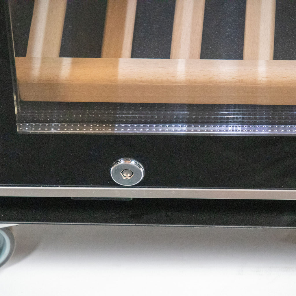 Nahaufnahme einer Glastür mit Schloss und Schlüsselloch unten. Hinter dem Glas befinden sich mehrere parallele Holzlamellen, typisch für einen Weinkühlschrank, wie den Weinkühlschrank 2 Zonen CASO WineComfort 38 in schwarz mit Fassholz-Umhausung. Die Konstruktion sieht aus wie ein Teil einer Lagereinheit oder eines Schranks auf Rädern, mit einem Metallrahmen, der das Glas und die Lamellen umgibt.