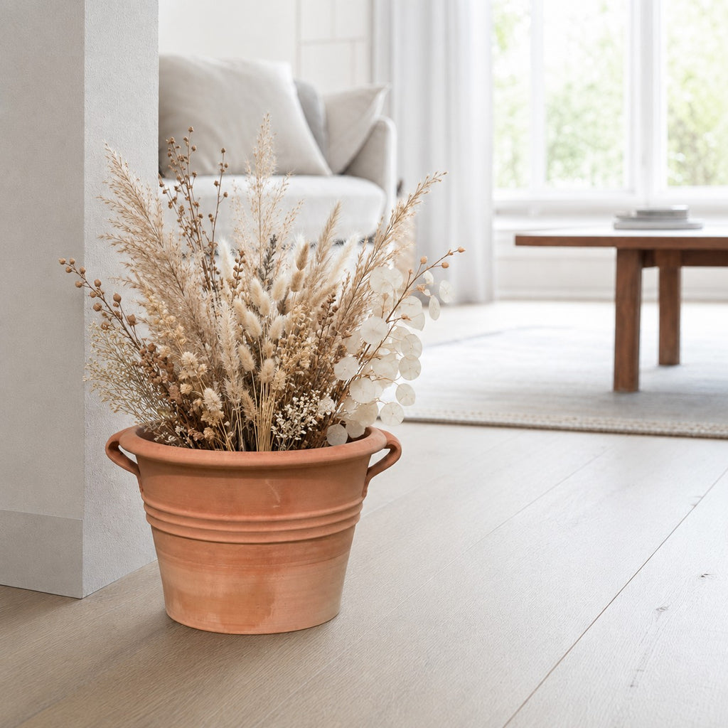 Ein Terrakotta-Pflanzkübel „Sorbus“ mit Henkeln (Durchmesser 40–60 cm), gefüllt mit getrockneten Gräsern und Blumen, steht auf einem hellen Holzboden in einem hellen, modernen Wohnzimmer mit neutraler Einrichtung.