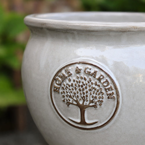Nahaufnahme eines hellgrauen Keramiktopfs mit einem eingeprägten runden Emblem, das einen Baum und die Worte "HOME & GARDEN" zeigt. Der Hintergrund besteht aus verschwommenem Grünzeug.
