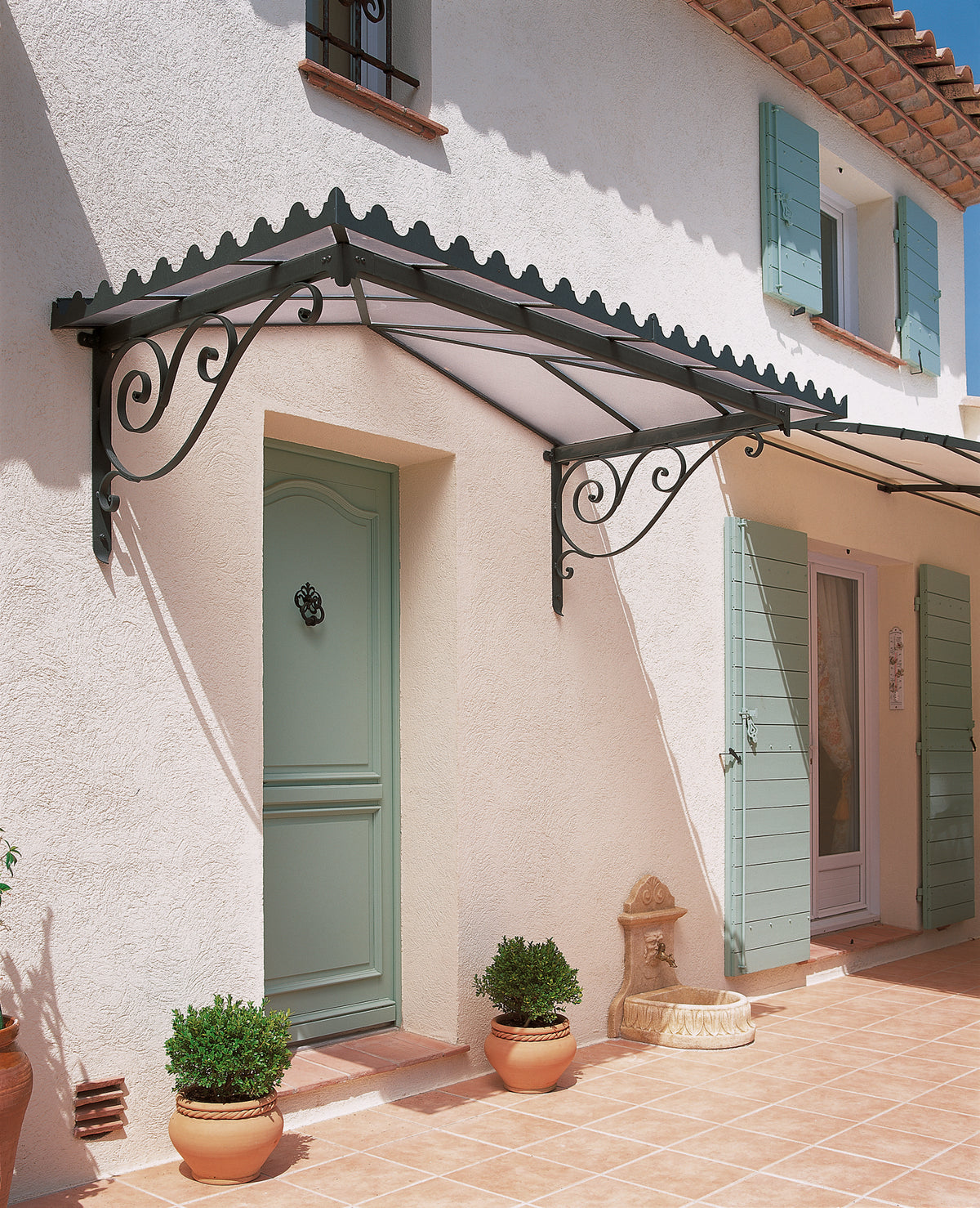 Außenansicht eines Hauses im französischen Landhausstil mit hellgrünen Fensterläden und Tür. Über der Tür erstreckt sich ein verziertes schwarzes Metall-Vordach mit Flachdach Dafne mit wirbelnden Mustern. Topfpflanzen stehen auf beiden Seiten der Tür und entlang des mit Terrakottafliesen ausgelegten Weges und verleihen dem Haus einen Hauch von Charme.