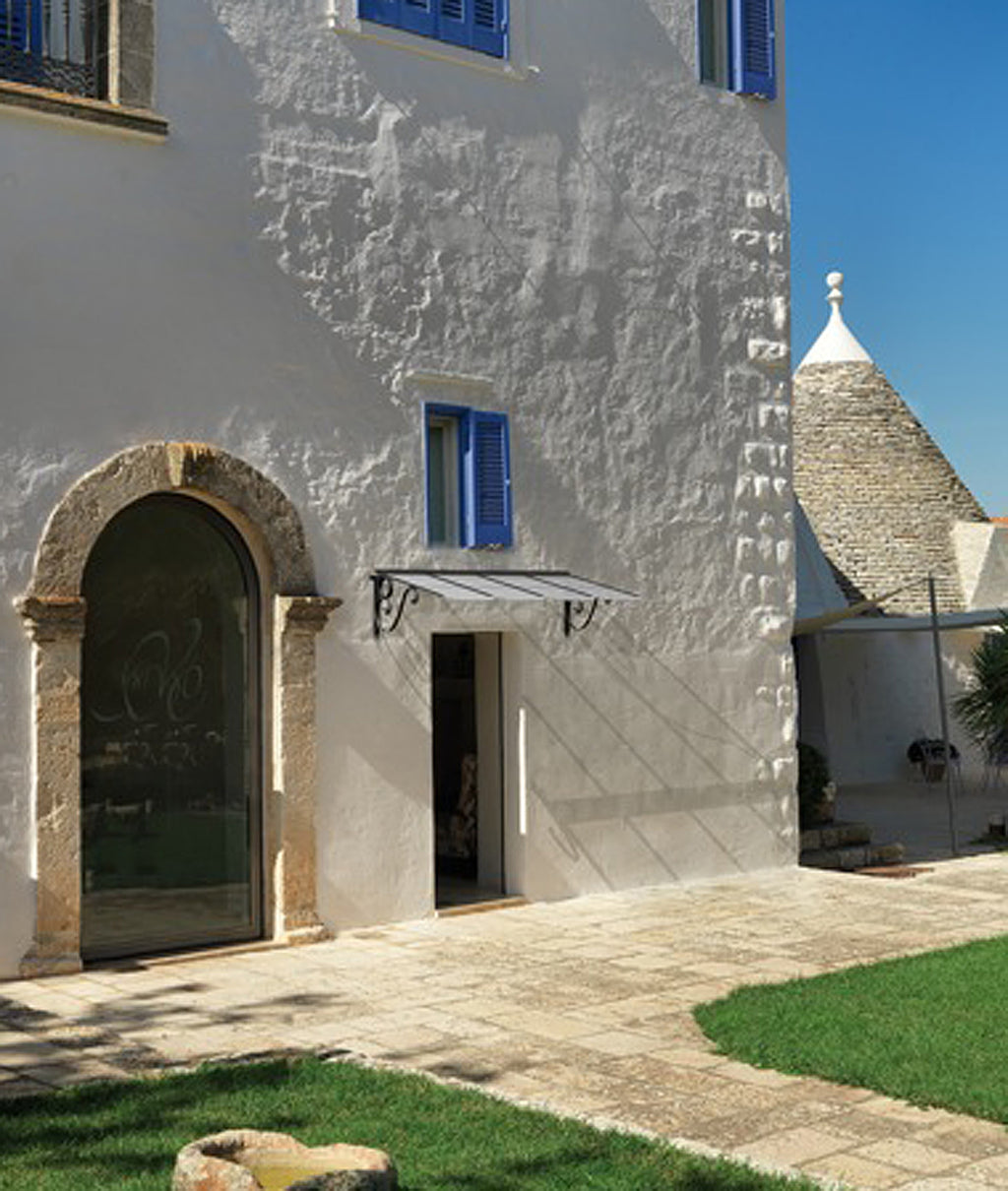 Ein weißes Gebäude im mediterranen Stil mit blauen Fensterläden und einem gewölbten Eingang öffnet sich zu einer Steinterrasse mit grünen Grasflächen. Im Hintergrund ist unter einem klaren blauen Himmel ein kleineres Gebäude mit einem konischen Dach zu sehen, ergänzt durch ein charmantes Vordach mit Flachdach über dem Eingang.