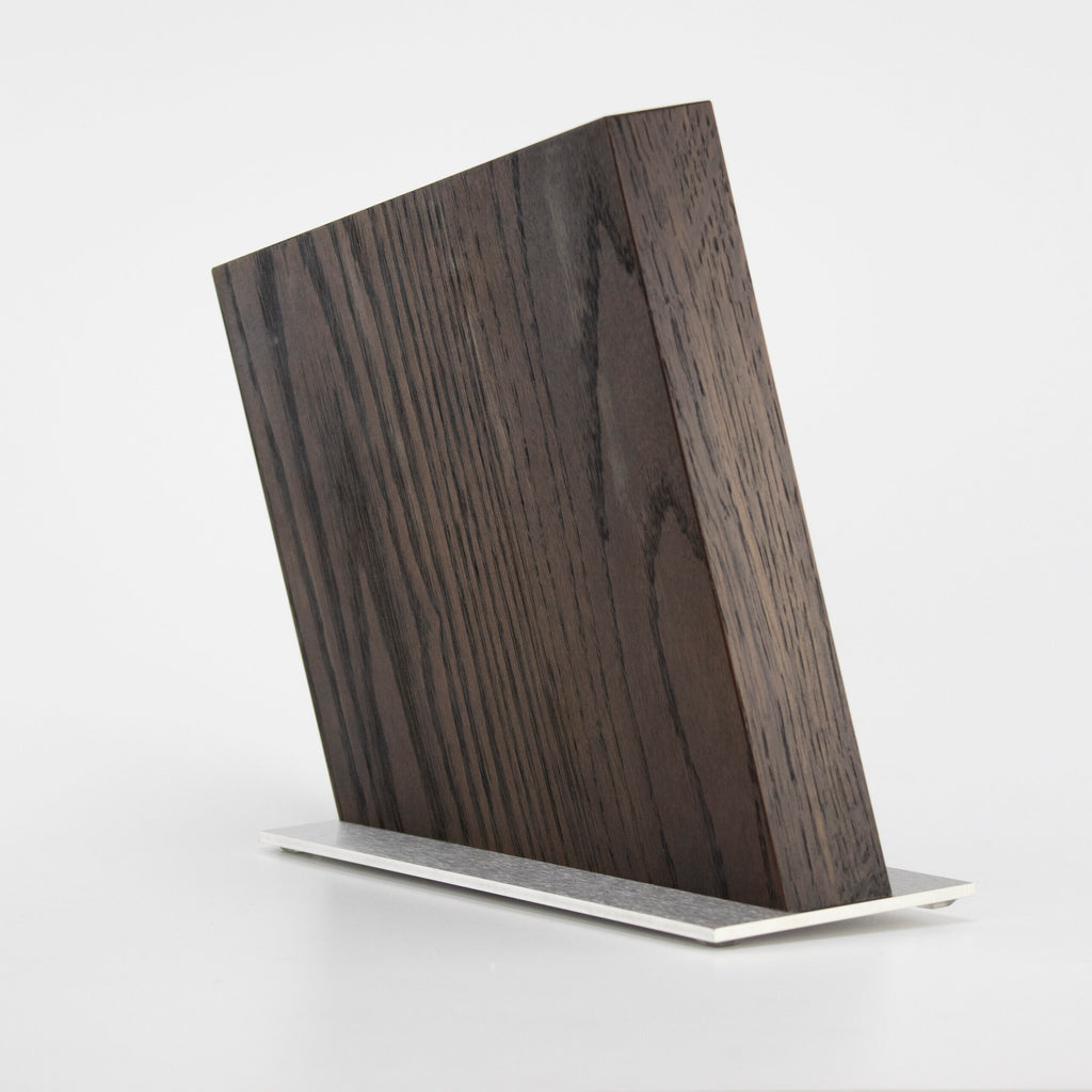 Ein dunkles, schräg geschnittenes Holzbrett steht aufrecht auf einem weißen Sockel. Das klare, minimalistische Design wird vor einem schlichten weißen Hintergrund präsentiert und hebt die Textur und Maserung des Holzes hervor – perfekt, um Ihr KAI Shun Classic White Damastmesser-Set + Magnet-Messerblock von Räuchereiche Böker in Szene zu setzen.