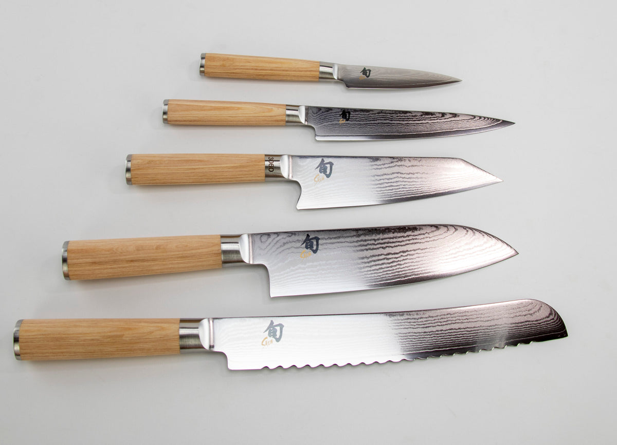 Ein Arrangement von fünf Küchenmessern aus dem KAI Shun Classic White Damastmesser-Set mit Holzgriffen, parallel ausgelegt auf einer weißen Oberfläche. Die Messer variieren in Größe und Form, darunter ein Schälmesser, Allzweckmesser, Kochmesser, Santokumesser und Brotmesser, jedes mit unterschiedlichem Klingendesign.