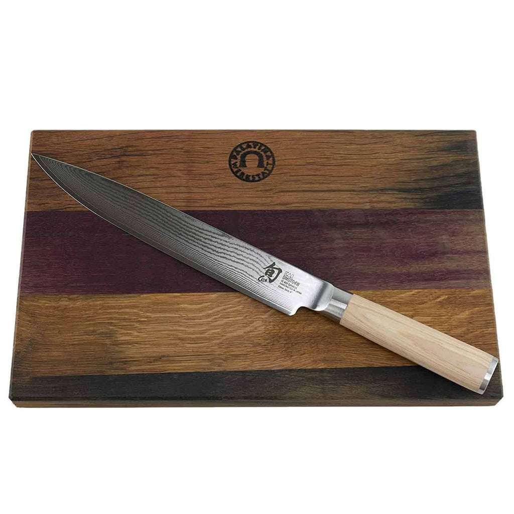 Ein Kai Shun Classic White DM-0704W Schinkenmesser mit einer polierten, dunkel gemusterten 23 cm Damastklinge und einem hellen Holzgriff liegt auf einem Fassholz Küchenbrett mit den Maßen 30x18 cm. Das Schneidebrett ist in dunklen und hellen Naturholztönen gehalten, mit leichten violetten Schattierungen akzentuiert und durch ein rundes Logo gekennzeichnet.