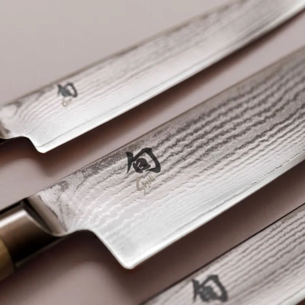 Das Kai Shun Classic White DM-0705W Brotmesser 23cm "THE MAGIC KAI WISHLIST" verfügt über eine Klinge aus Damaszenerstahl mit eingravierten Kanji, die diagonal auf einer hellen Oberfläche angezeigt werden, um das komplizierte Wellenmuster hervorzuheben.