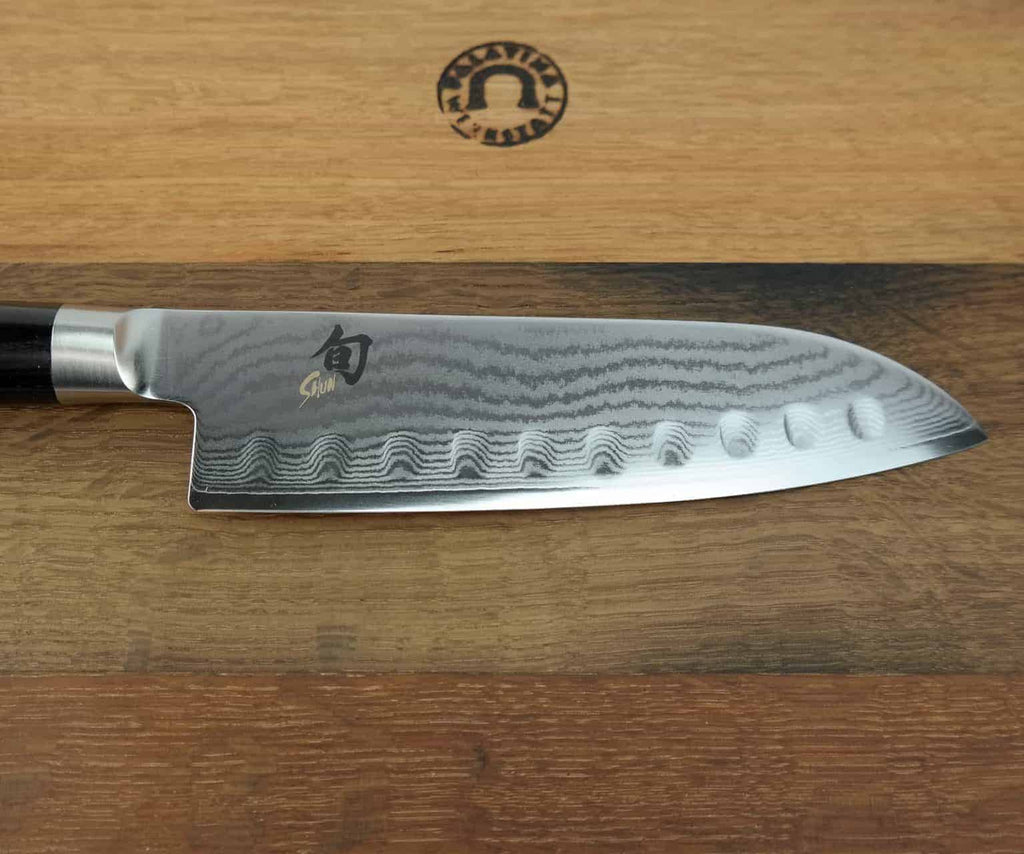 Eine Nahaufnahme des Kai Shun Classic Santoku DM-0718-Messers mit einer 18 cm langen Klinge mit Damastmuster und gewellten Einkerbungen entlang der Schneide. Die glänzende Klinge trägt ein Logo und einen Text, und der dunkle Griff ergänzt das elegante Design. Das Messer ruht auf einem Fassholzbrett mit den Maßen 34 x 21 cm, auf dem sich ein Markenemblem befindet.