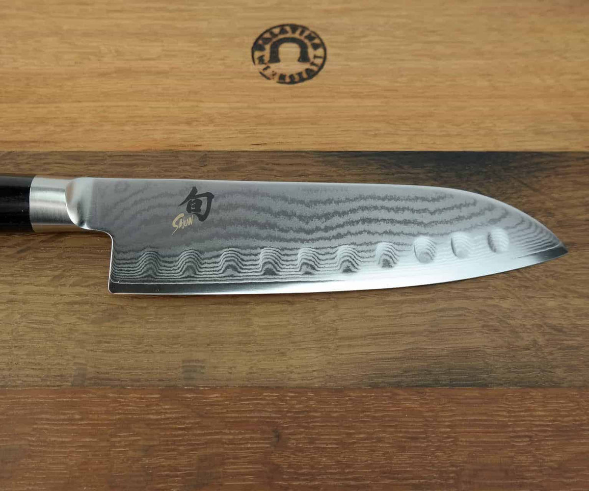 Eine Nahaufnahme des Kai Shun Classic Santoku DM-0718-Messers mit einer 18 cm langen Klinge mit Damastmuster und gewellten Einkerbungen entlang der Schneide. Die glänzende Klinge trägt ein Logo und einen Text, und der dunkle Griff ergänzt das elegante Design. Das Messer ruht auf einem Fassholzbrett mit den Maßen 34 x 21 cm, auf dem sich ein Markenemblem befindet.