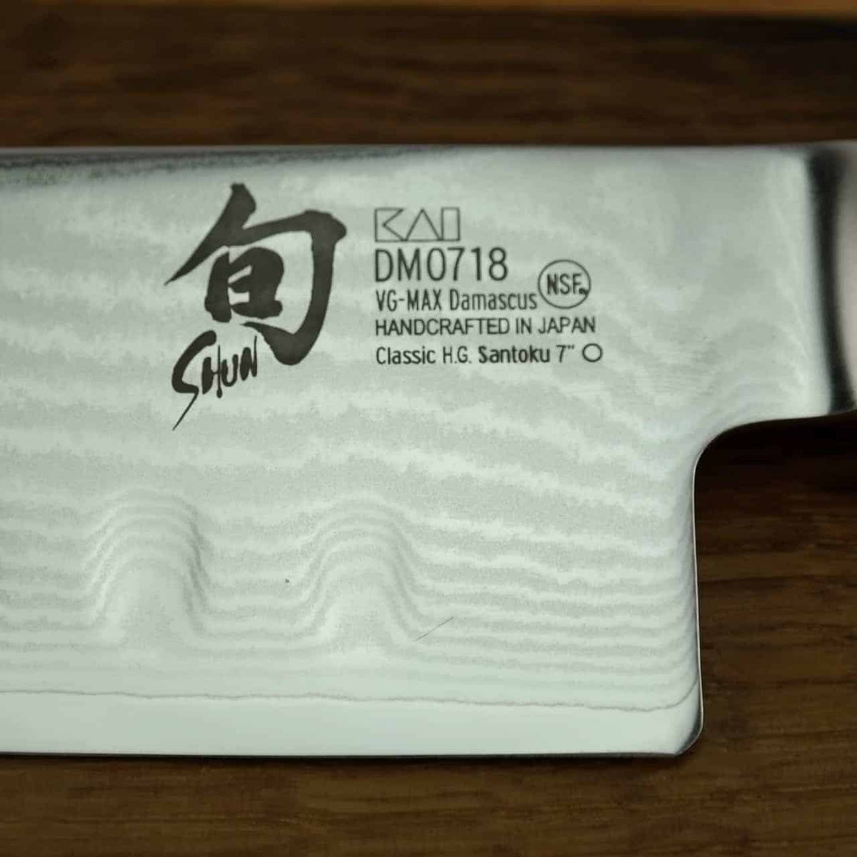 Eine Nahaufnahme der Klinge des Kai Shun Classic Santoku zeigt Inschriften, darunter ein japanisches Schriftzeichen, „KAI“, „DM0718“, „VG-MAX Damascus“, „HANDCRAFTED IN JAPAN“, „NSF“ und „Classic H.G. Santoku“. Die reflektierende 18 cm lange Klinge besticht durch ein atemberaubendes Damaszenermuster und einen präzisionssteigernden Kullenschliff.
