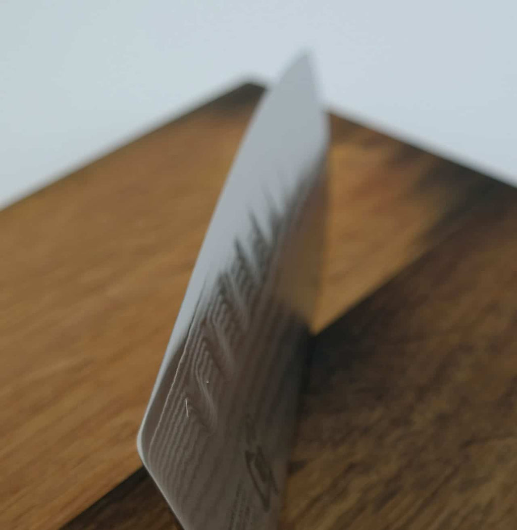 Ein Kai Shun Classic Santoku DM-0718 mit Kullenschliff (18 cm Klinge) liegt hochkant auf einem Fassholzbrett (34 x 21 cm) vor einem schlichten Hintergrund. Auf der Oberfläche der Klinge sind schwache, unleserliche Texte und Symbole zu sehen. Das Bild konzentriert sich auf die Schneide des Santokumessers und bietet eine Nahaufnahme.