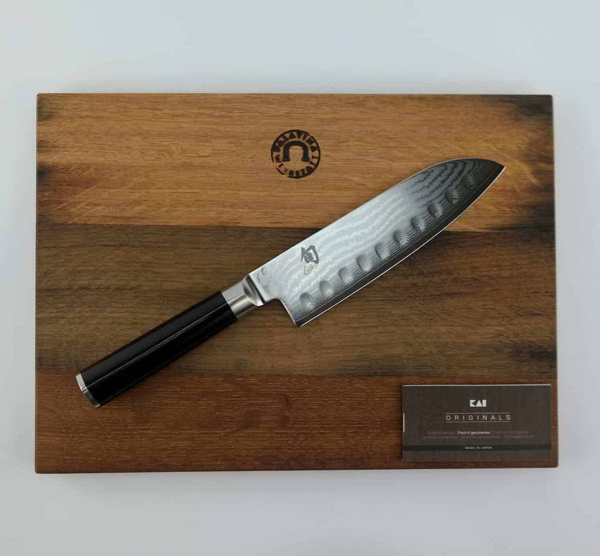 Ein Kai Shun Classic Santoku DM-0718 mit Kullenschliff, 18 cm Klinge Kochmesser mit polierter, gemusterter Klinge und schwarzem Griff ruht auf einem Fassholzbrett mit den Maßen 34x21cm. Unten rechts auf dem Schneidebrett befindet sich eine kleine Karte mit dem Markennamen „Kai“.