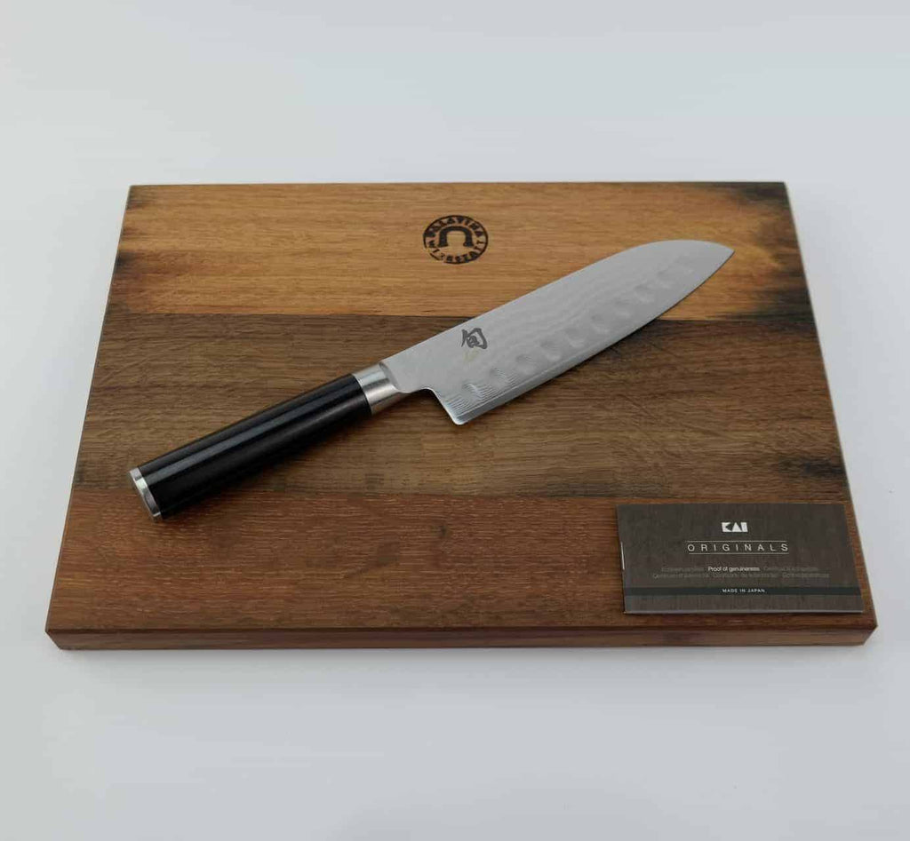 Ein hochwertiges Kai Shun Classic Santoku DM-0718 Kochmesser mit 18 cm Kullenschliff-Klinge und schwarzem Griff liegt auf einem 34x21 cm großen Fassholzbrett-Schneidebrett. In der unteren rechten Ecke des Schneidebretts ist eine schwarz-silberne Plakette mit dem Markenlogo und „Originals“ angebracht.