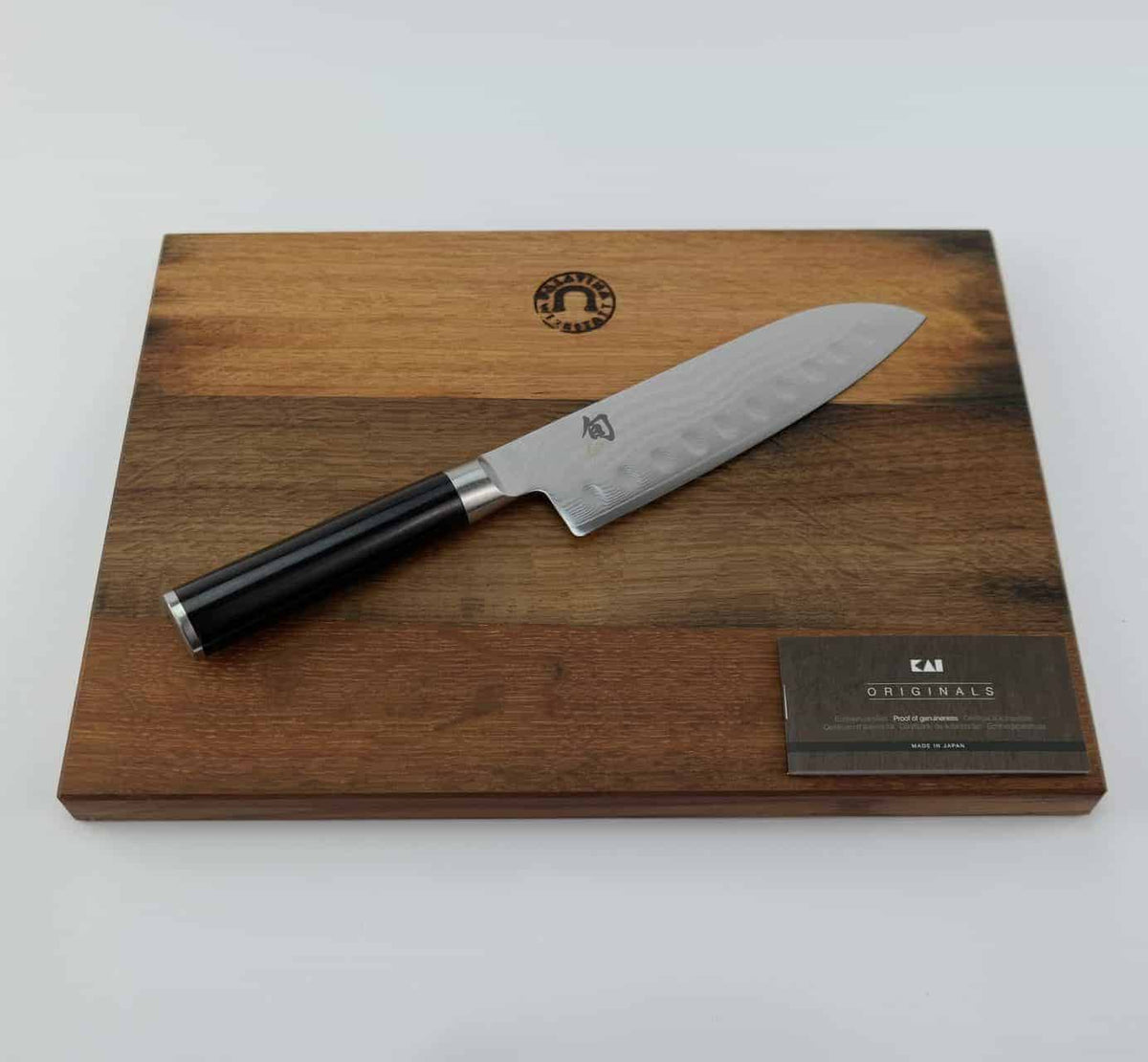 Ein hochwertiges Kai Shun Classic Santoku DM-0718 Kochmesser mit 18 cm Kullenschliff-Klinge und schwarzem Griff liegt auf einem 34x21 cm großen Fassholzbrett-Schneidebrett. In der unteren rechten Ecke des Schneidebretts ist eine schwarz-silberne Plakette mit dem Markenlogo und „Originals“ angebracht.