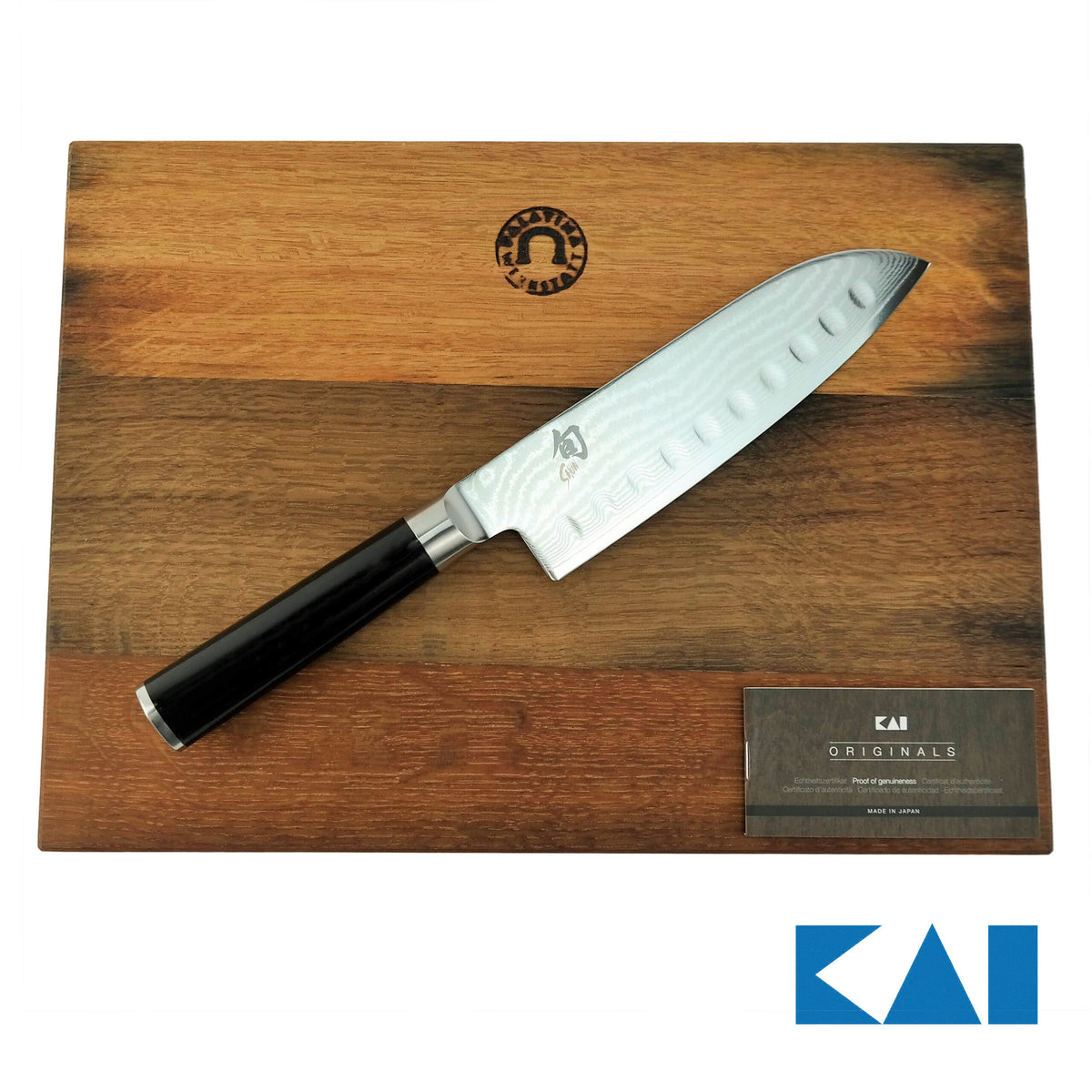 Ein Küchenmesser aus rostfreiem Stahl, das Kai Shun Classic Santoku DM-0718 mit Kullenschliff, mit schwarzem Griff liegt auf einem Fassholzbrett mit den Maßen 34x21 cm. Die 18 cm lange Klinge des Messers weist ein Wellenmuster und mehrere Grübchen auf, die als Kullenschliff bezeichnet werden. Neben dem Messer liegt eine kleine Karte, auf der in der unteren rechten Ecke des Bildes das KAI-Logo zu sehen ist.