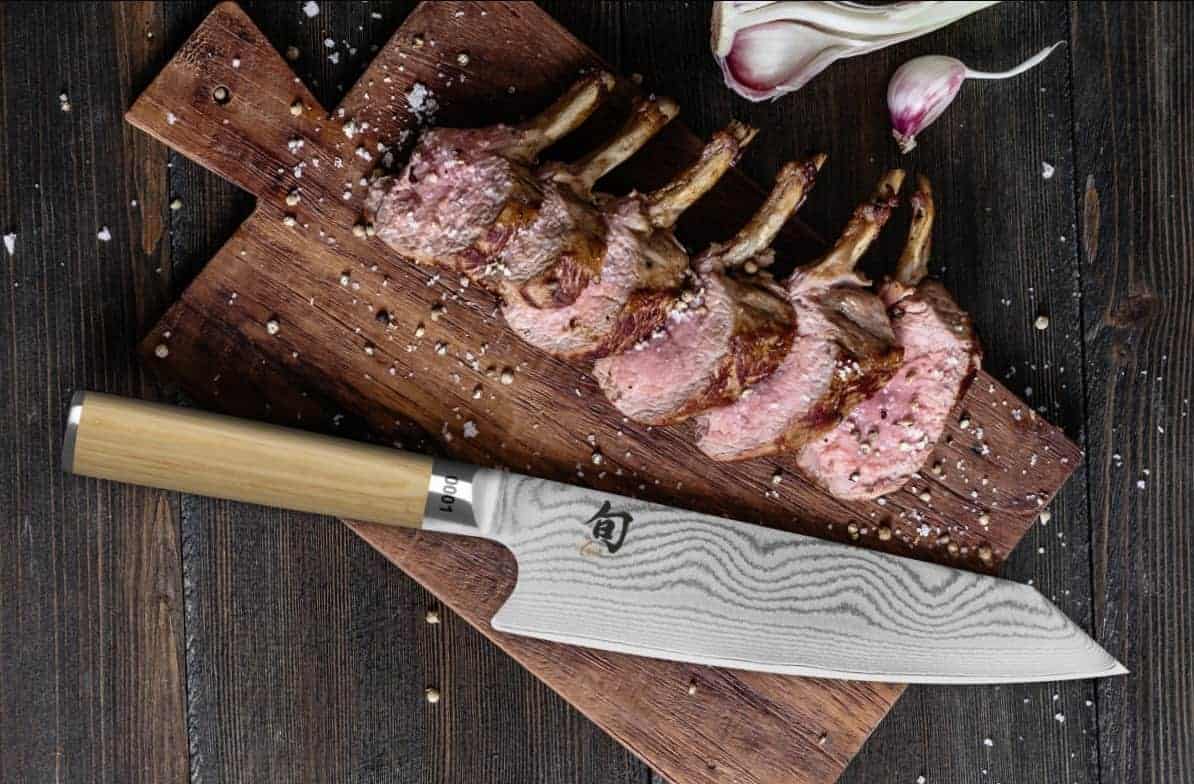 Auf einem hölzernen Schneidebrett liegen mehrere gekochte Lammkoteletts, garniert mit grobem Salz. Neben den Koteletts liegt ein japanisches Kochmesser mit Holzgriff. Auf dem Brett, das auf einem dunklen Holztisch steht, sind Knoblauchzehen verstreut.