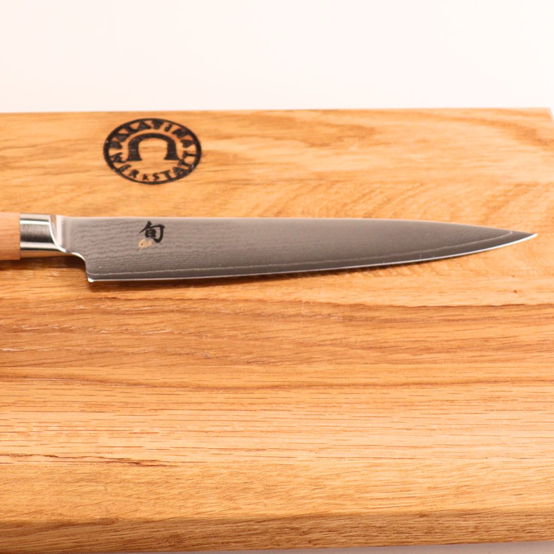 Das Kai Shun Classic White DM-0701W Allzweckmesser (15 cm VG10 Klinge) mit Holzgriff ruht auf dem mitgelieferten 30x20 cm großen Weinfass Küchenbrett, das mit einem dezenten Klingenmuster, Kanji in der Nähe des Griffs und einem runden Logo auf dem Brett versehen ist.