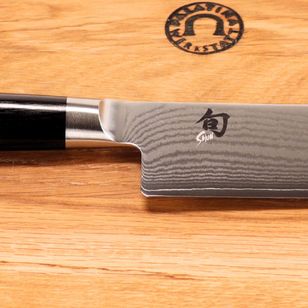 Eine Nahaufnahme des Kai Shun Classic Nakiri Messersets - DM-0728 16,5 cm Damast (32 Lagen) mit dunklem Griff, der auf dem mitgelieferten 30x20 cm großen Eichenholz-Schneidebrett mit eingraviertem kreisförmigen Logo ruht.
