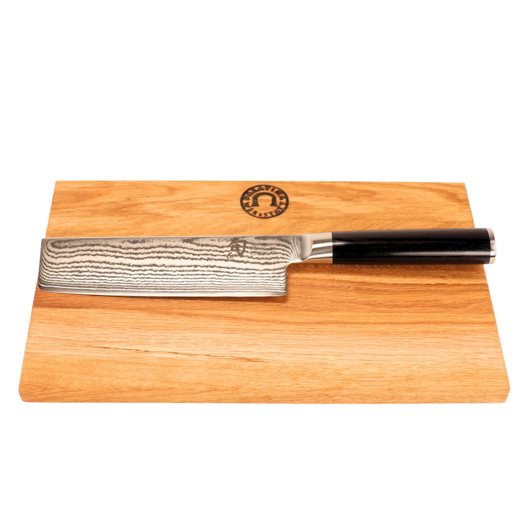 Das Kai Shun Classic Nakiri Messer Set (DM-0728, 16,5 cm, 32-lagiger Damast) mit schwarzem Griff wird mit einem 30x20 cm großen Schneidebrett aus Eichenholz geliefert, das ein dunkles, kreisförmiges Logo in der oberen Mitte trägt.
