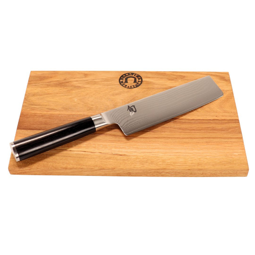 Das Kai Shun Classic Nakiri Messer Set (DM-0728, 16,5 cm, 32-lagiger Damast) besteht aus einem silbernen Messer mit schwarzem Griff, das auf einem 30 x 20 cm großen, rechteckigen Schneidebrett aus Eichenholz mit einem runden Logo am oberen Rand liegt.